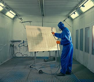 Pracownik w niebieskim kombinezonie ochronnym lakierujący duży biały panel w profesjonalnej kabinie lakierniczej wyposażonej w system wentylacji i oświetlenia.