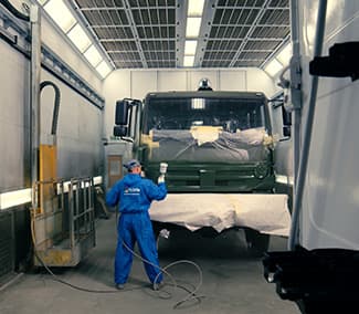 Kabina lakiernicza do pojazdów ciężarowych z pracownikiem w niebieskim kombinezonie wykonującym prace lakiernicze. Widoczny samochód ciężarowy w trakcie procesu lakierowania.